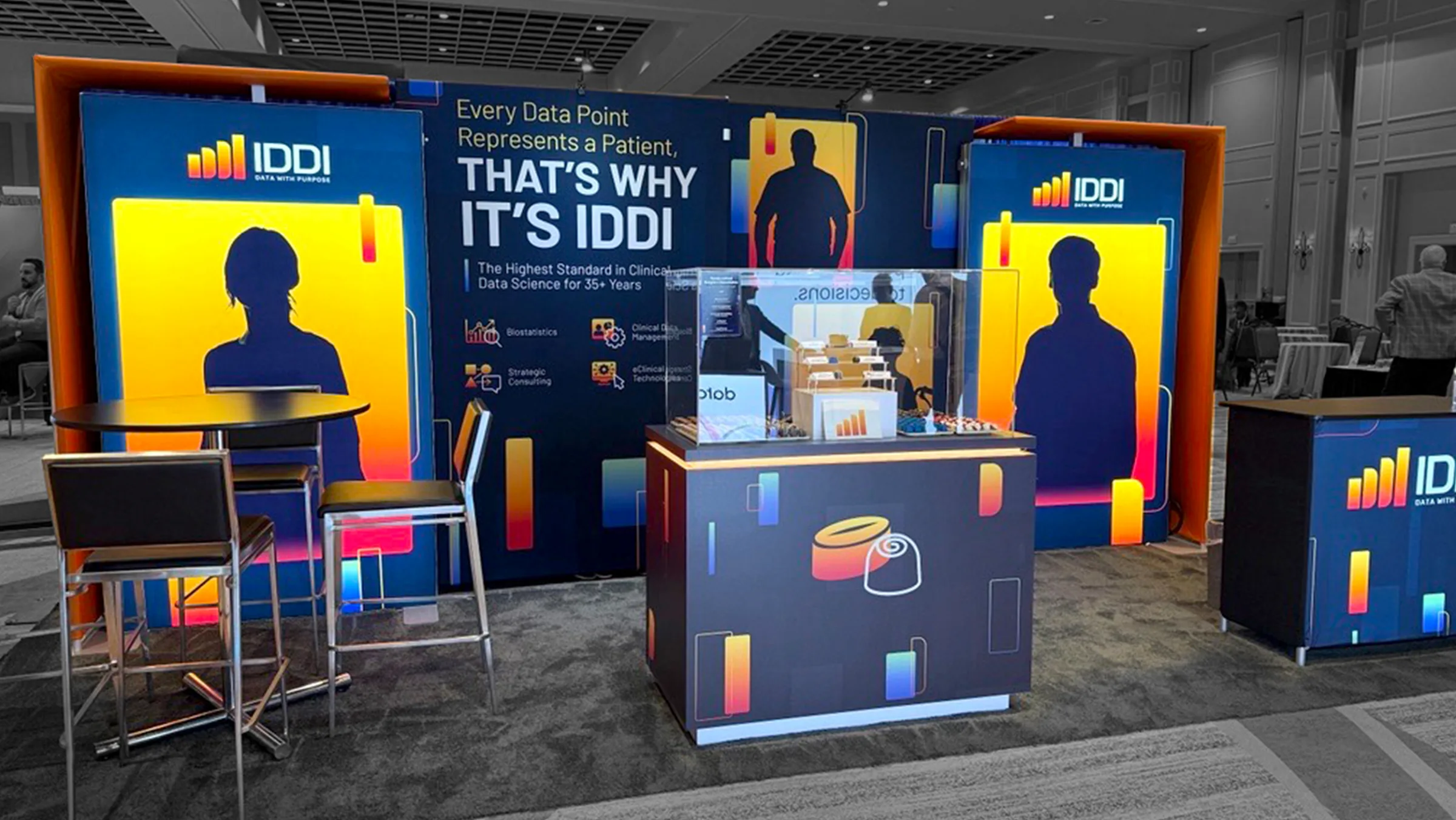 IDDI trade show booth featuring bold branded backdrops with the tagline "That's Why It's IDDI," silhouette graphics, a reception desk, and seating area.