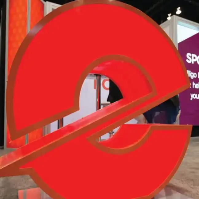 A large red three-dimensional Elligo lowercase "e" logo sculpture on the floor of a busy trade show exhibition hall.