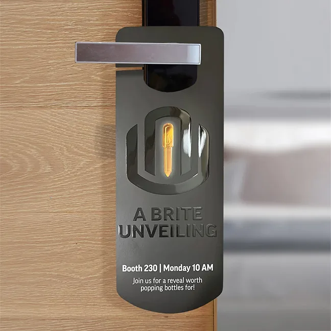 Dark gray hotel door hanger promoting "A Brite Unveiling" event at Booth 230 on Monday at 10 AM, featuring a glowing key logo.